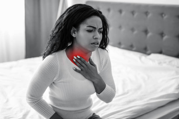 Throat Pain Concept. Sad sick young African American woman coughing, suffering from sore throat, sitting in bed at home. Ill lady touching neck with hand, red highlight area, black and white