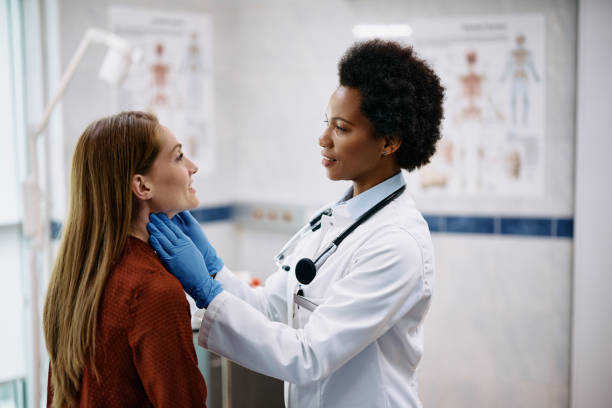 African American female doctor touching woman's neck during thyroid exam at medical clinic.